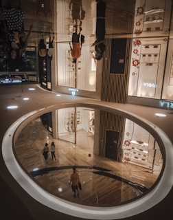 An indoor shopping mall with a shiny reflective floor creating upside-down reflections of people walking. Stores have bright displays, and floral patterns decorate some sections. The floor has a circular opening that reveals a lower level.
