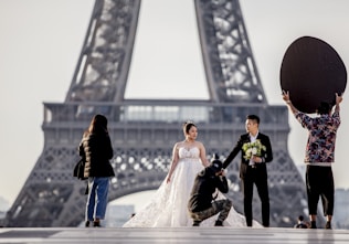 A couple dressed in wedding attire poses near the Eiffel Tower, with the bride in a white dress holding a bouquet and the groom in a dark suit. A photographer kneels to capture the moment while another person holds a large reflector to enhance the lighting. Another individual stands to the side, possibly assisting.