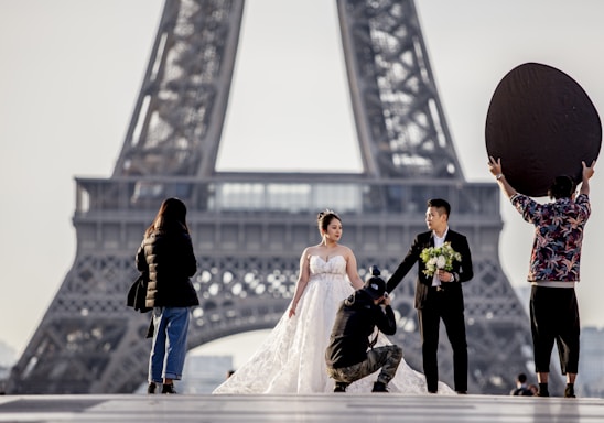 A couple dressed in wedding attire poses near the Eiffel Tower, with the bride in a white dress holding a bouquet and the groom in a dark suit. A photographer kneels to capture the moment while another person holds a large reflector to enhance the lighting. Another individual stands to the side, possibly assisting.