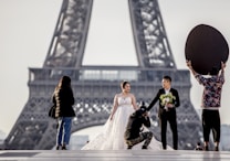 A couple dressed in wedding attire poses near the Eiffel Tower, with the bride in a white dress holding a bouquet and the groom in a dark suit. A photographer kneels to capture the moment while another person holds a large reflector to enhance the lighting. Another individual stands to the side, possibly assisting.