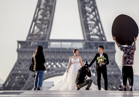 A couple dressed in wedding attire poses near the Eiffel Tower, with the bride in a white dress holding a bouquet and the groom in a dark suit. A photographer kneels to capture the moment while another person holds a large reflector to enhance the lighting. Another individual stands to the side, possibly assisting.