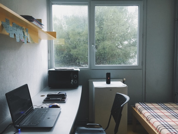 A tidy room with a minimalist setup includes a desk with a laptop, a mouse, and some books or notepads. Above the desk, there's a shelf with sticky notes and additional materials. To the right, there is a bed with a plaid-patterned bedspread. A small refrigerator sits beneath a window, and a speaker is placed on top of it. The room appears to be well-lit by the natural light coming from the window, which shows a view of green trees outside.