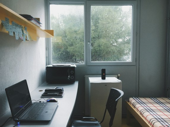 A tidy room with a minimalist setup includes a desk with a laptop, a mouse, and some books or notepads. Above the desk, there's a shelf with sticky notes and additional materials. To the right, there is a bed with a plaid-patterned bedspread. A small refrigerator sits beneath a window, and a speaker is placed on top of it. The room appears to be well-lit by the natural light coming from the window, which shows a view of green trees outside.
