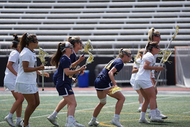 A group of women in athletic gear are playing lacrosse on a sports field. The players are wearing protective eyewear and holding lacrosse sticks. Some are dressed in blue uniforms, while others are in white. The field has a turf surface, and rows of empty bleachers are visible in the background.
