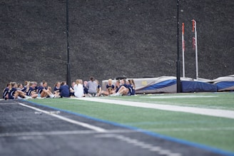 A group of people, likely a sports team, is sitting together on a field. They are wearing matching athletic uniforms, suggesting team unity, and seem to be engaged in a discussion or meeting. In the background, there is sports equipment and a high fence.