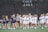 A group of female lacrosse players from two different teams is on a sports field. One team is wearing dark blue uniforms while the other is in white. The players are interacting, possibly after a game, with some shaking hands or patting each other on the back.