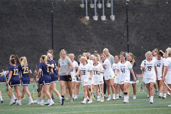 A group of female lacrosse players from two different teams is on a sports field. One team is wearing dark blue uniforms while the other is in white. The players are interacting, possibly after a game, with some shaking hands or patting each other on the back.