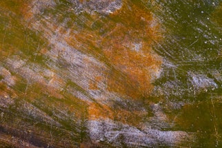 A textured surface featuring a mix of earthy tones, primarily olive green with patches of rusty orange and hints of white. The surface appears weathered, with visible scratches and a rough finish, suggesting it could be a metal or wooden material.