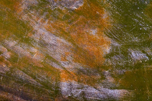 A textured surface featuring a mix of earthy tones, primarily olive green with patches of rusty orange and hints of white. The surface appears weathered, with visible scratches and a rough finish, suggesting it could be a metal or wooden material.