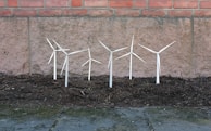 Several small wind turbine models are planted in a patch of soil against a stone wall with bricks at the top. These miniature turbines appear to be made of white or light-colored material.