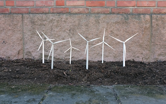 Several small wind turbine models are planted in a patch of soil against a stone wall with bricks at the top. These miniature turbines appear to be made of white or light-colored material.