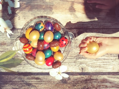 A collection of vibrantly colored, decorated eggs are placed in a clear glass bowl on a rustic wooden surface. Nearby, a hand is holding a single golden egg. Sunlight casts shadows across the scene, adding warmth and depth. White blossoms and leaves decorate the edges, enhancing the festive, springtime atmosphere.