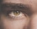 A close-up of a human eye with greenish-brown iris color, showing detailed textures of the skin and eyebrow hair. The lighting emphasizes the natural patterns and colors within the eye.