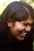 A person with long dark hair, wearing a stud earring, is laughing joyfully. Their eyes are closed, and their facial expression conveys happiness and amusement.