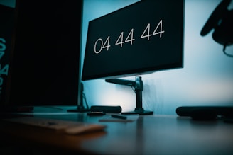 A digital clock display on a monitor reads 04:44:44. The setup includes a desk with peripheral devices, a mouse, and a keyboard, against a blue-tinted background. The lighting is dim and the environment suggests a tech or office space.