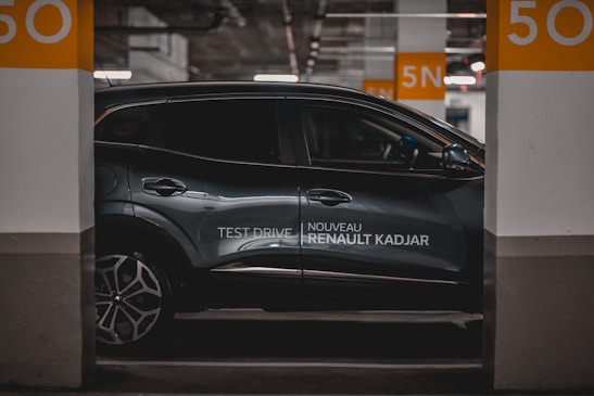A sleek, modern car is parked in an indoor parking space. The vehicle is metallic gray with a glossy finish and features alloy wheels. It is positioned between two concrete pillars, marked with the number 50 in large orange font. The branding 'Nouveau Renault Kadjar' and 'Test Drive' is visible on the side of the car. The surrounding area is dimly lit, contributing to a sophisticated atmosphere.