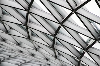 A close-up view of an architectural structure featuring a grid-like pattern of intersecting metal beams forming geometric shapes. The beams are painted in shades of gray and white, with glass panels in between allowing natural light to filter through.