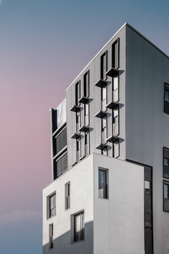 A modern, multi-story building with a sleek design featuring vertical lines and multiple rectangular windows. The structure combines light gray and darker gray tones, with clean, geometric shapes. The sky in the background is a gradient of soft blue and pink hues, adding a serene ambiance.