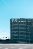 A multi-story parking garage with a modern glass facade is visible. The large letter 'P8' is prominently displayed on the building, indicating it is part of a series of parking garages. The structure has a series of horizontal lines suggesting the floors of the parking facility. Surrounding the building, there are several street lamps and traffic signs, and the sky is clear with a vibrant blue color.
