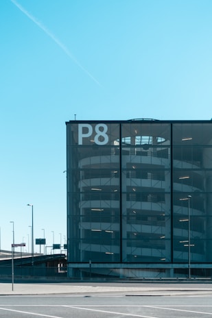 A multi-story parking garage with a modern glass facade is visible. The large letter 'P8' is prominently displayed on the building, indicating it is part of a series of parking garages. The structure has a series of horizontal lines suggesting the floors of the parking facility. Surrounding the building, there are several street lamps and traffic signs, and the sky is clear with a vibrant blue color.