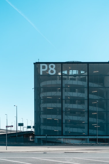 A multi-story parking garage with a modern glass facade is visible. The large letter 'P8' is prominently displayed on the building, indicating it is part of a series of parking garages. The structure has a series of horizontal lines suggesting the floors of the parking facility. Surrounding the building, there are several street lamps and traffic signs, and the sky is clear with a vibrant blue color.