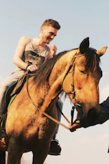 A smiling instructor helping a young rider adjust their posture on a calm horse in a sunlit arena.
