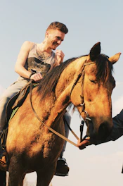 A smiling instructor guiding a young rider on a gentle horse along a sunlit trail.