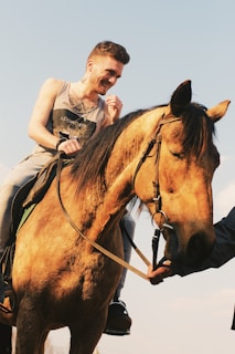 A person is sitting on a horse while smiling and looking down. The horse has a brown coat and the person is wearing casual clothes with a tank top. Another person's hand is visible, gently holding the horse's bridle.