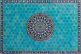 A detailed Islamic geometric pattern featuring a central circular design with intricate interlocking shapes crafted from blue, white, and green colors. This is set against a background of hexagonal turquoise tiles, bordered by a smaller repeating triangular pattern.