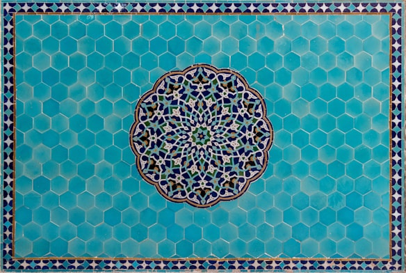 A detailed Islamic geometric pattern featuring a central circular design with intricate interlocking shapes crafted from blue, white, and green colors. This is set against a background of hexagonal turquoise tiles, bordered by a smaller repeating triangular pattern.