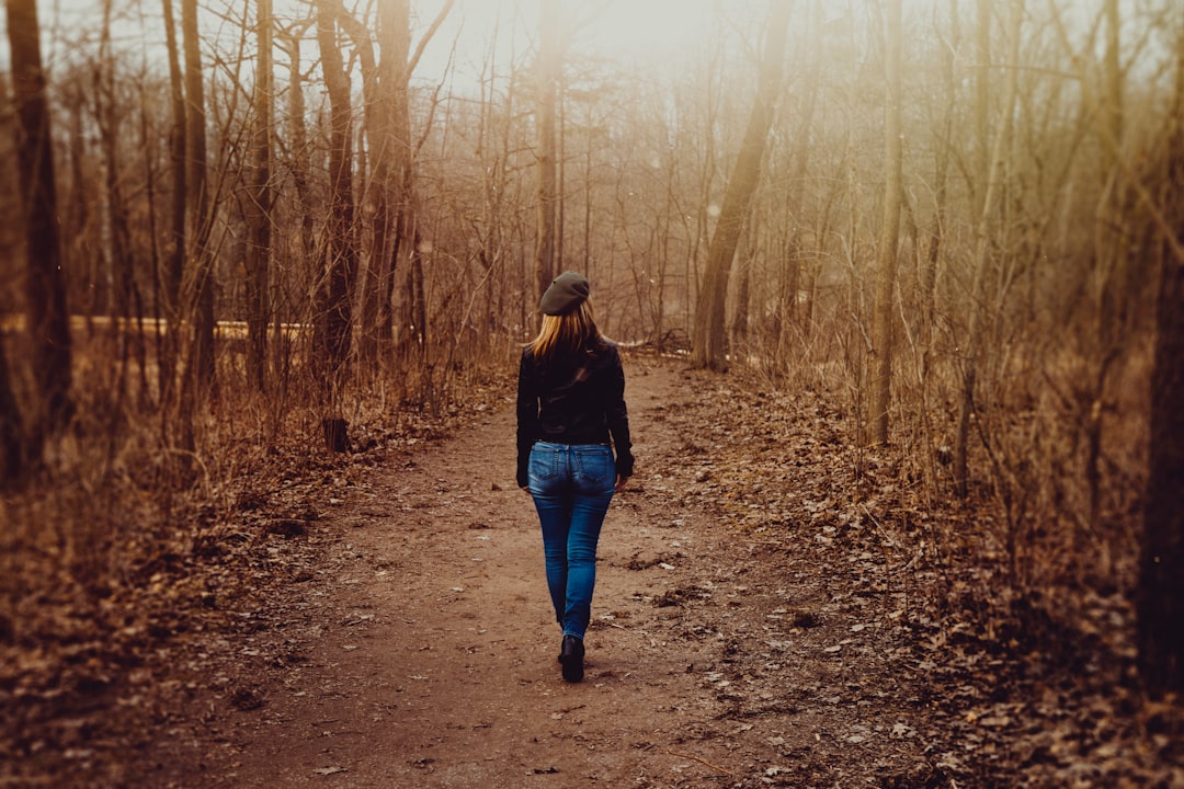 woman in black sweater and blue denim skinny jeans walks on dirt road beside leafless trees