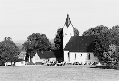 A peaceful rural scene featuring a small church with a steeple surrounded by trees and a grassy field. People appear to be gathered outside the building, sitting on benches or chairs in a relaxed setting.