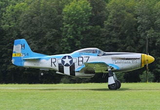 A vintage military aircraft with the words 'Louisiana Kid' painted on its side, parked on a grassy field. The plane features a blue tail fin, black-and-white stripes, and a star emblem. The surrounding background is a forest with dense green trees.