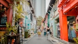 A vibrant street scene featuring colorful buildings adorned with various decorations such as plants and lanterns. People are casually strolling along the narrow street, exploring shops and cafes. Signs and boards are visible, indicating business names and promotions.