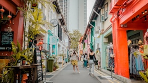 A vibrant street scene featuring colorful buildings adorned with various decorations such as plants and lanterns. People are casually strolling along the narrow street, exploring shops and cafes. Signs and boards are visible, indicating business names and promotions.
