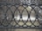 A decorative black wrought iron fence with an intricate design consisting of interlocking circles and ornate elements. Behind the fence, sparse dry bushes and a small paved path are visible. The scene appears to be in an outdoor urban setting.