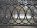 A decorative black wrought iron fence with an intricate design consisting of interlocking circles and ornate elements. Behind the fence, sparse dry bushes and a small paved path are visible. The scene appears to be in an outdoor urban setting.