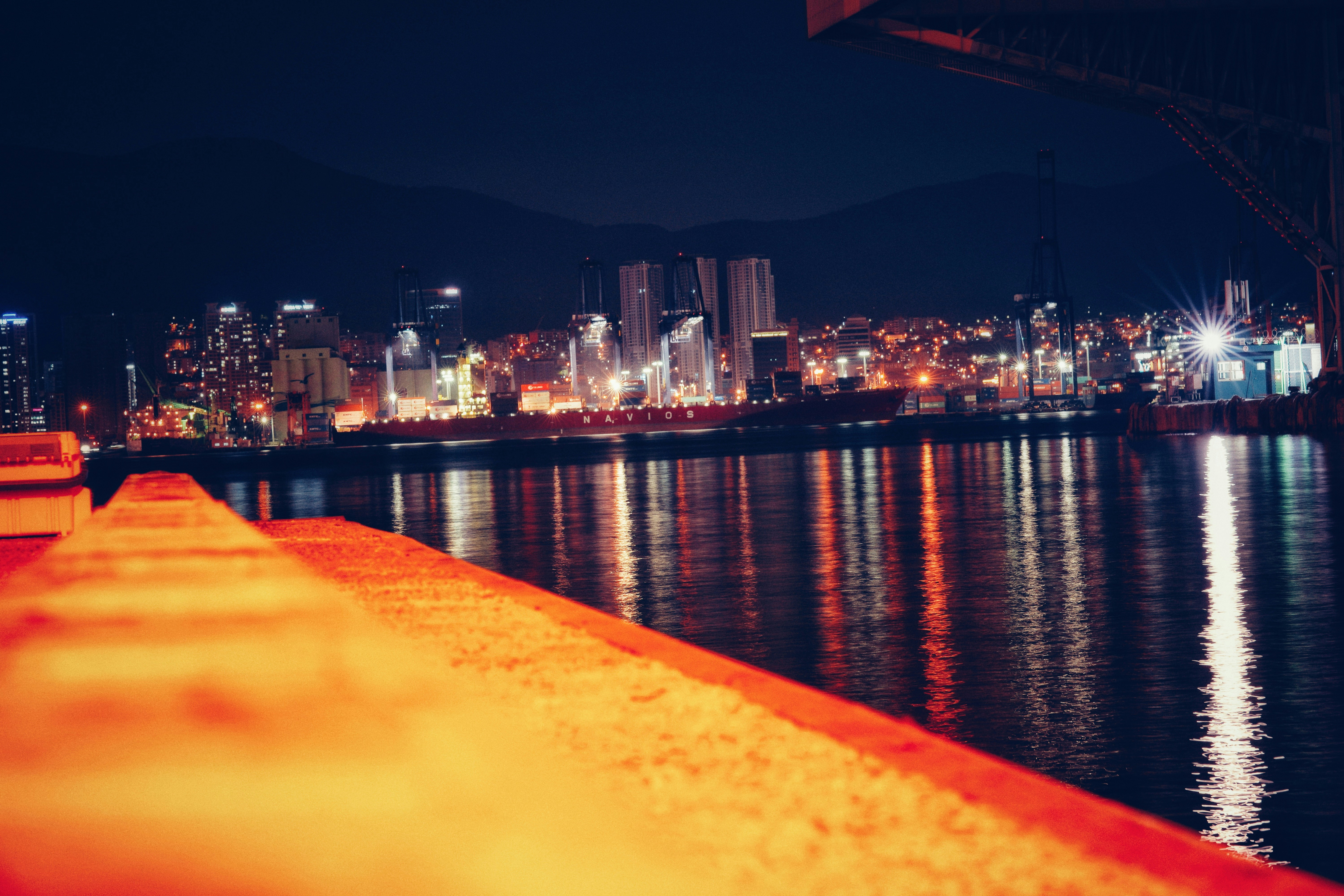 Large cargo ship at night with bright port lights reflecting on dark water, industrial shipping terminal in background