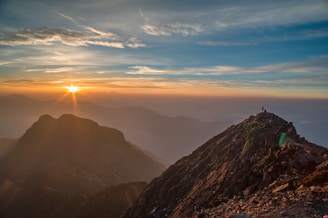 A breathtaking sunrise over a rugged mountain range with a solitary figure standing on the peak, gazing into the horizon. The sky is filled with vibrant hues of orange and blue, while the sunlight casts a warm glow across the landscape, highlighting the textures of the rocky terrain.
