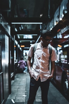 A young person stands on a dimly lit city street, wearing a white button-up shirt with a backpack. They are next to a red double-decker bus with reflections on its windows. The setting is urban, possibly at night, with people in the background.