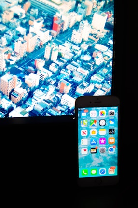 A smartphone with various app icons is placed in front of a screen displaying an aerial view of a densely packed cityscape with numerous buildings and streets.