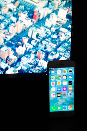 A smartphone with various app icons is placed in front of a screen displaying an aerial view of a densely packed cityscape with numerous buildings and streets.