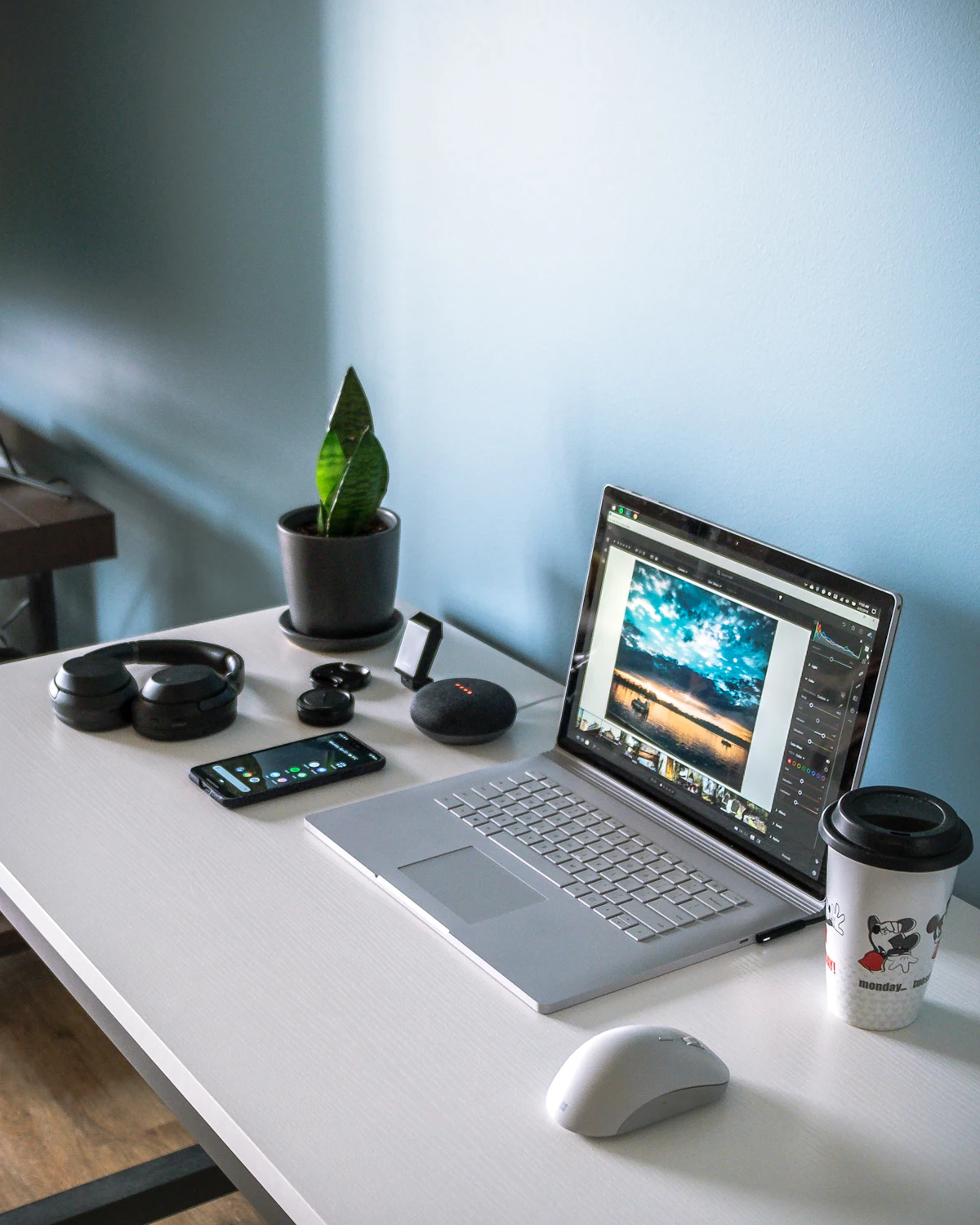 Un portable sur un bureau blanc devant un mur bleu clair, un logiciel de retouche affichant une photo de nuages à l'écran, et à côté un casque, une sansevière, un téléphone et une enceinte connectée