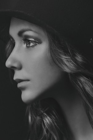 A grayscale portrait of a woman in profile, wearing a hat. Her long hair is styled in soft waves, and her facial features are elegantly highlighted with subtle lighting.
