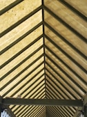 A symmetrical view of a roof structure with a series of evenly spaced triangular beams converging to form a perspective. The roof panels are a translucent material that allows light to filter through, creating a warm glow.