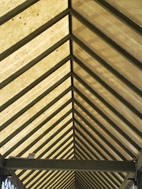A symmetrical view of a roof structure with a series of evenly spaced triangular beams converging to form a perspective. The roof panels are a translucent material that allows light to filter through, creating a warm glow.