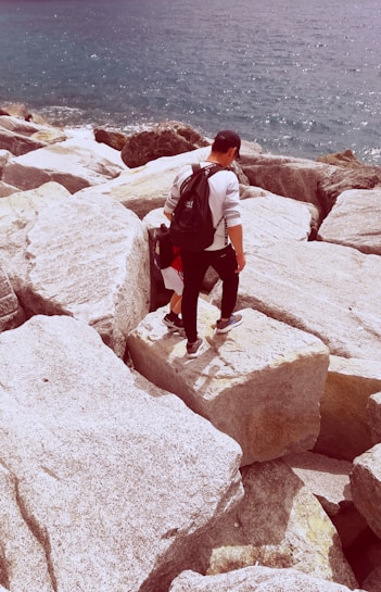 A person wearing a backpack and cap is walking across large, weathered rocks next to a body of water. The waves gently lap against the shore, creating a sense of movement. The scene suggests a coastal environment with a focus on the rugged terrain.