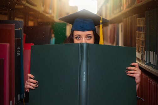 A person wearing a graduation cap peers over a large green book while standing in a library aisle. The shelves are filled with variously colored books, and sunlight filters through the top of the shelves, creating a warm ambiance.