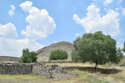 A large ancient pyramid surrounded by scattered trees and stone structures under a bright blue sky with fluffy white clouds. The scene includes grassy areas and a path leading up the pyramid.