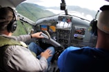 Two people are seated in the cockpit of a small aircraft, wearing headsets. The person on the left is operating the controls while the other observes. The cockpit features various instruments, dials, and screens. A view of forested mountains and a body of water is visible through the windshield.